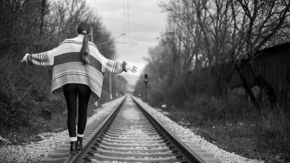 A woman in a sweater balances on a railway track, captured in a moody black and white setting.